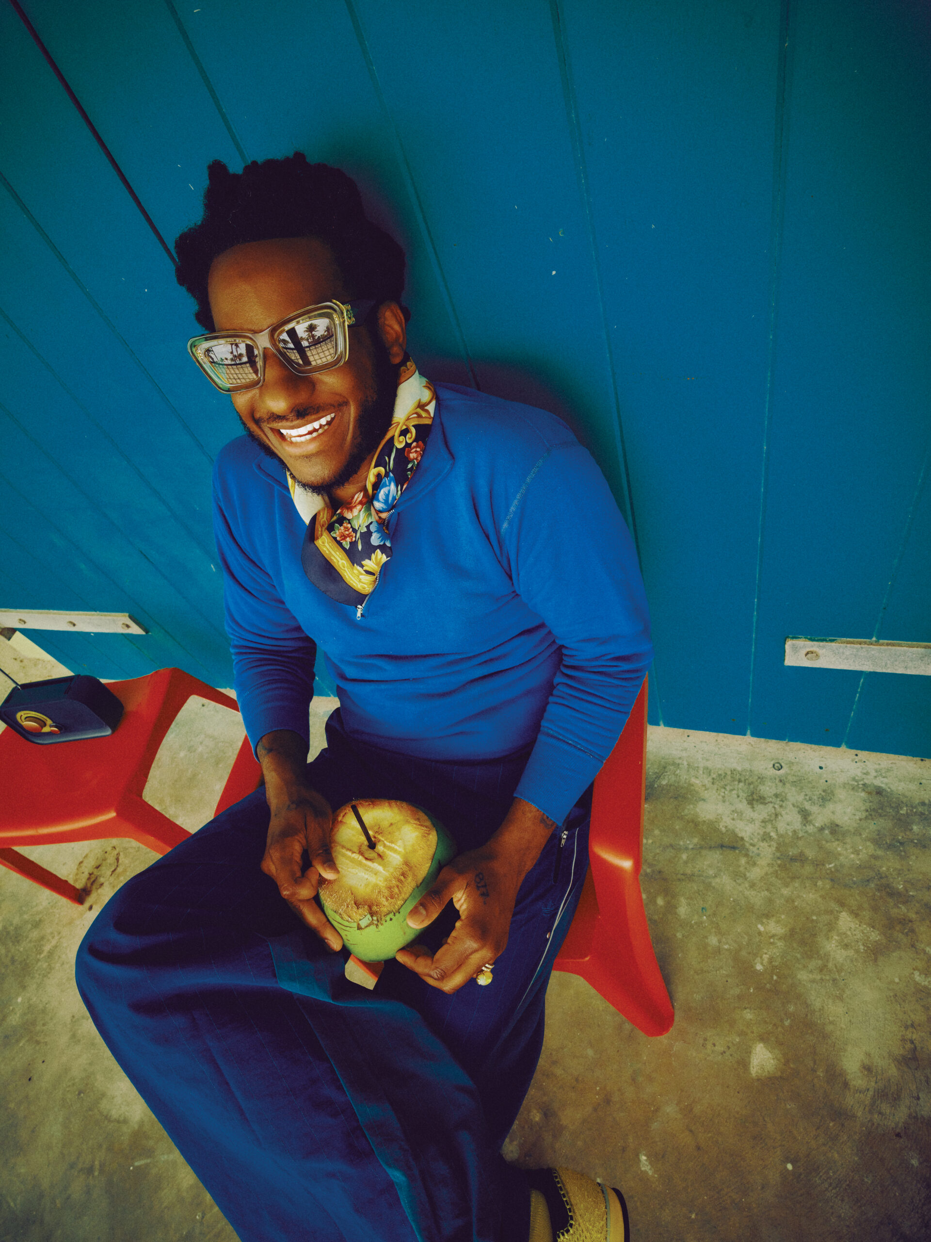 Leon Bridges smiling in sunglasses and blue clothing, seated on a red chair holding a coconut.